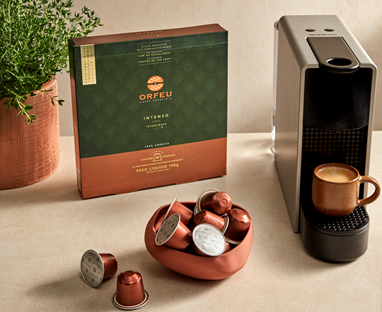 Pacote de cápsulas de café Orfeu Intenso ao lado de uma máquina de café com uma xícara de café. Cápsulas de café estão dispostas em uma tigela e na mesa.