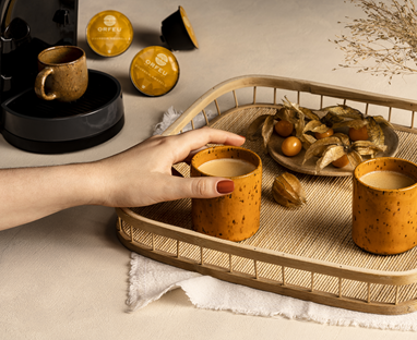 Bandeja com xícaras de café, frutas e cápsulas de café Orfeu. Uma mão está segurando uma xícara de café próximo a uma máquina de café.